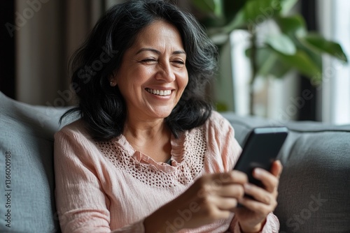 Smiling Mexican mature woman making call seated on sofa, hold smartphone, browsing information, chatting with family through messenger, enjoy digital communication and pastime on internet. Modern tech