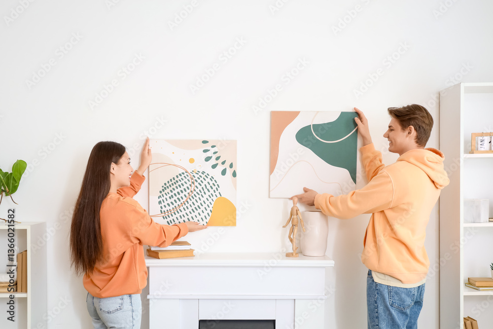 Fototapeta premium Young couple hanging paintings on wall above fireplace at home