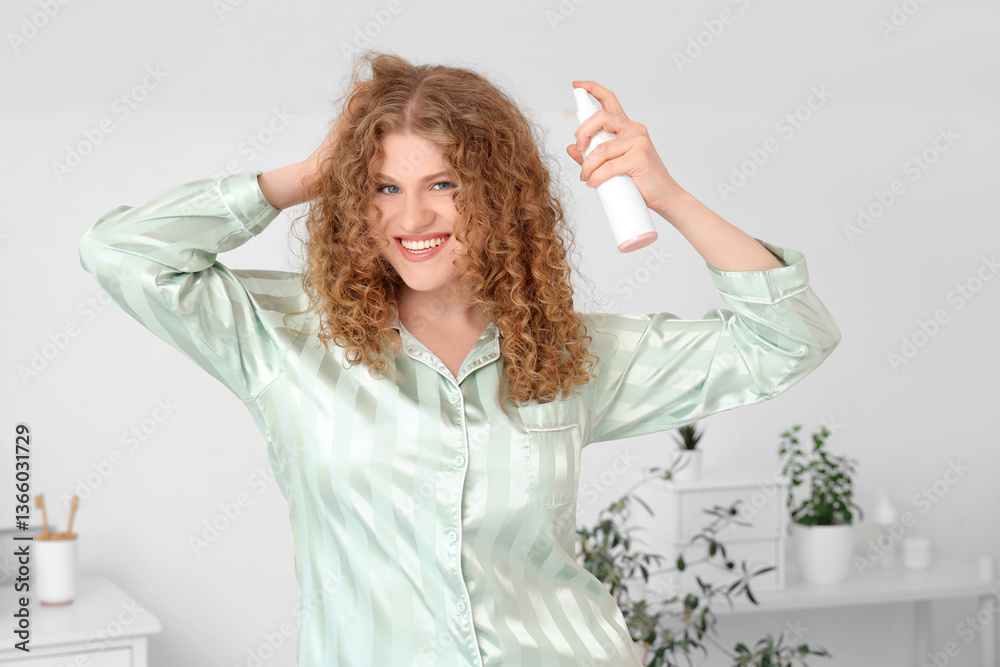 Naklejka premium Smiling young woman with curly hair applying hair spray for styling and care in pajamas, she is wearing pajamas, performing her morning beauty routine at home