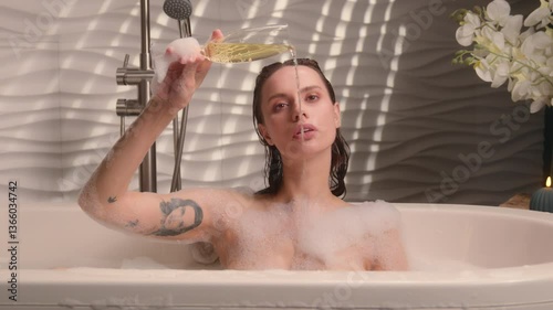 Naked Caucasian girl female sexy woman beautiful relaxed lady relaxing in bathroom foam rest in bathtub enjoying foamy beauty routine spa pleasure holding glass alcohol pouring out champagne in water