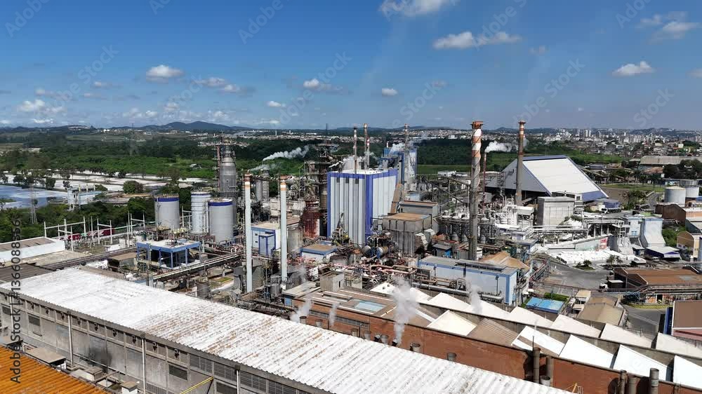Vídeo aéreo em 4K de uma indústria de papel em plena produção durante o dia. Imagens claras mostram chaminés, galpões, maquinário e atividade industrial sob céu azul e ensolarado