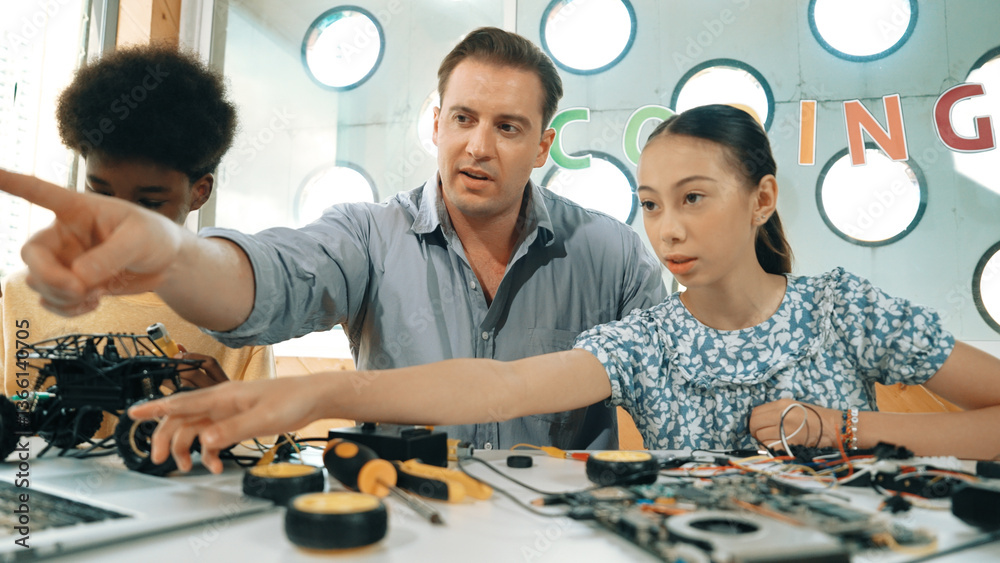 Caucasian student asking teacher about code and pointing at laptop screen. Professional instructor answer girl question while boy repair car model at table with electronic tool placed. Edification.