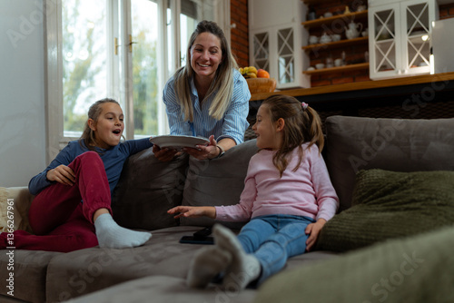 Happy mother serving a plate of delicious food to her two daughters, who are relaxing together on the cozy sofa in their inviting living room, enjoying quality time at home