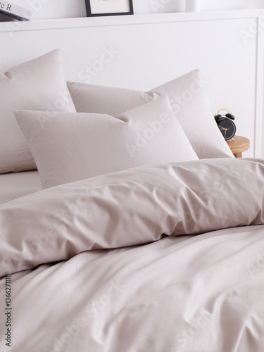 Bedroom with a bed, fluffy down comforter, three pillows, wooden bedside table and alarm clock. Close-up side view vertical photo