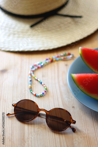 Plate with watermelon slices, glass of orange juice or cocktail, straw hat, colorful beaded necklace and sunglasses on the wooden table. Summer essentials concept. Selective focus.