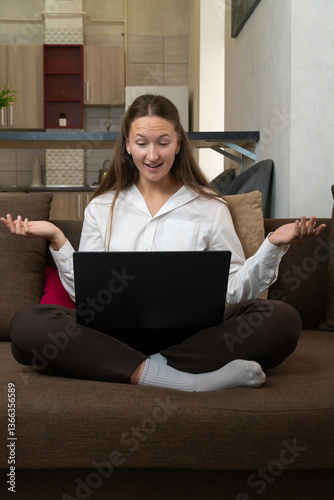 Businesswoman sitting on sofa with laptop on her lap and looking surprised while working from home, she's raising her arms and opening her mouth in shock. Vertical photo