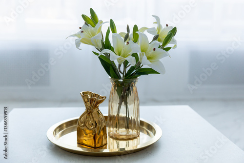 Bouquet of white artificial lilies in a golden vase on a table on a light background.