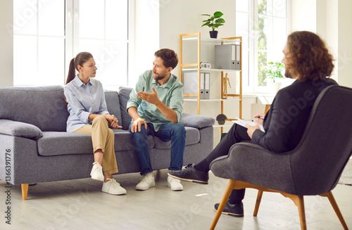 Obraz na płótnie Young family couple sitting on sofa at therapist office, talking to each other about their relationship problems. Professional psychologist providing support to husband and wife during therapy session w salonie