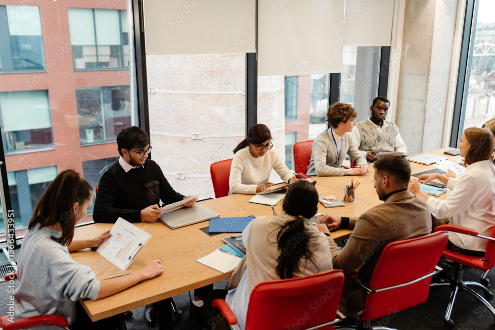 A multinational group of young people engaging in a productive discussion during their internship at a modern office, working on laptops and documents in a bright meeting room with large windows.
