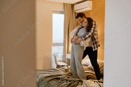 Konstfotografi A pregnant woman and her partner stand in a cozy bedroom, embracing warmly