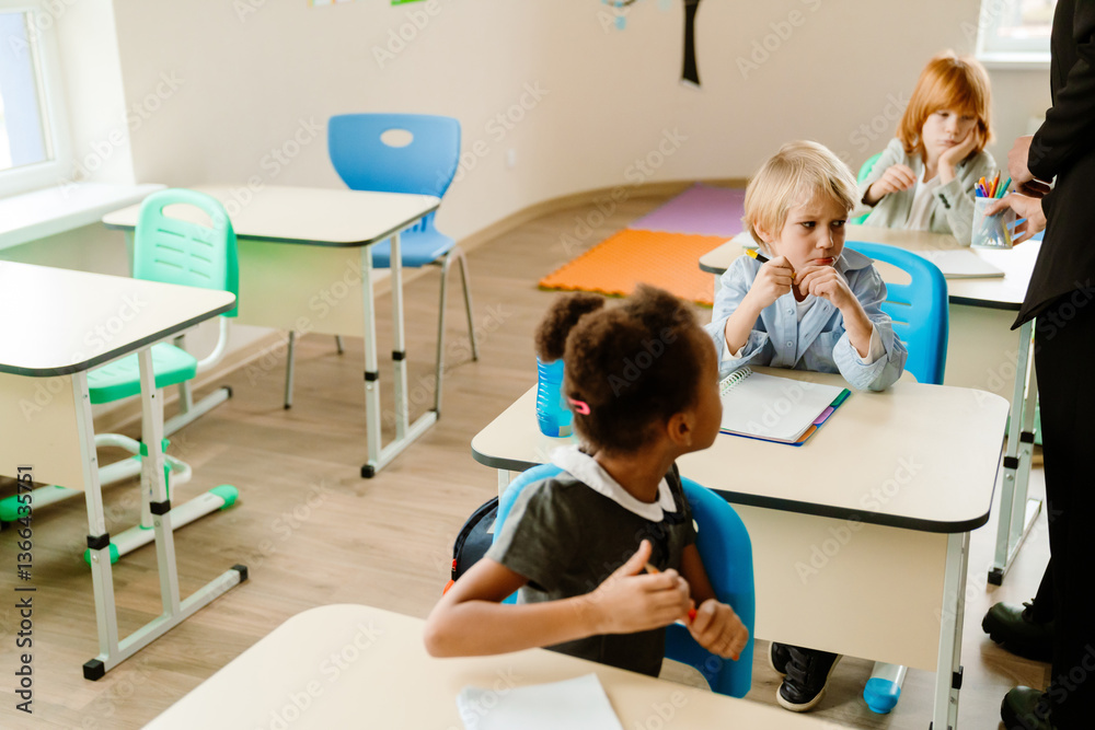 Obraz premium A group of children sit at their desks in a bright classroom. A blonde boy in a light blue shirt holds a pencil while looking focused, while a Black girl in uniform is engaged in an activity.