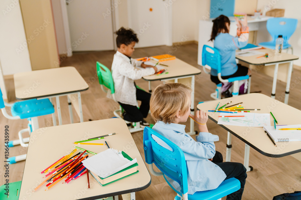 Obraz premium boy in a light blue shirt sits at his desk, holding a pencil while looking at his drawing. Other students are focused on their creative activities