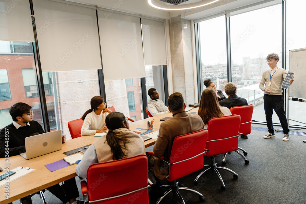 Fototapeta premium A group of eight employees sit at a table and look at a male intern standing next to them and smiling while holding a clipboard, in an office, during an internship