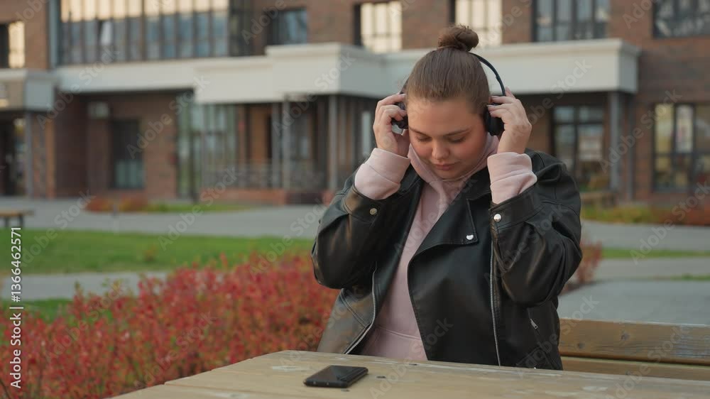 White woman seated outdoors puts headset on and reaches for phone on table with red flowers swaying in wind and modern office building in background during calm afternoon moment