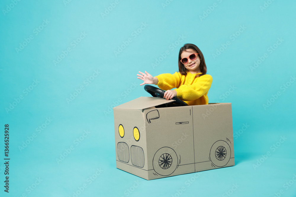 Little girl waving while driving car made with cardboard on light blue background. Space for text