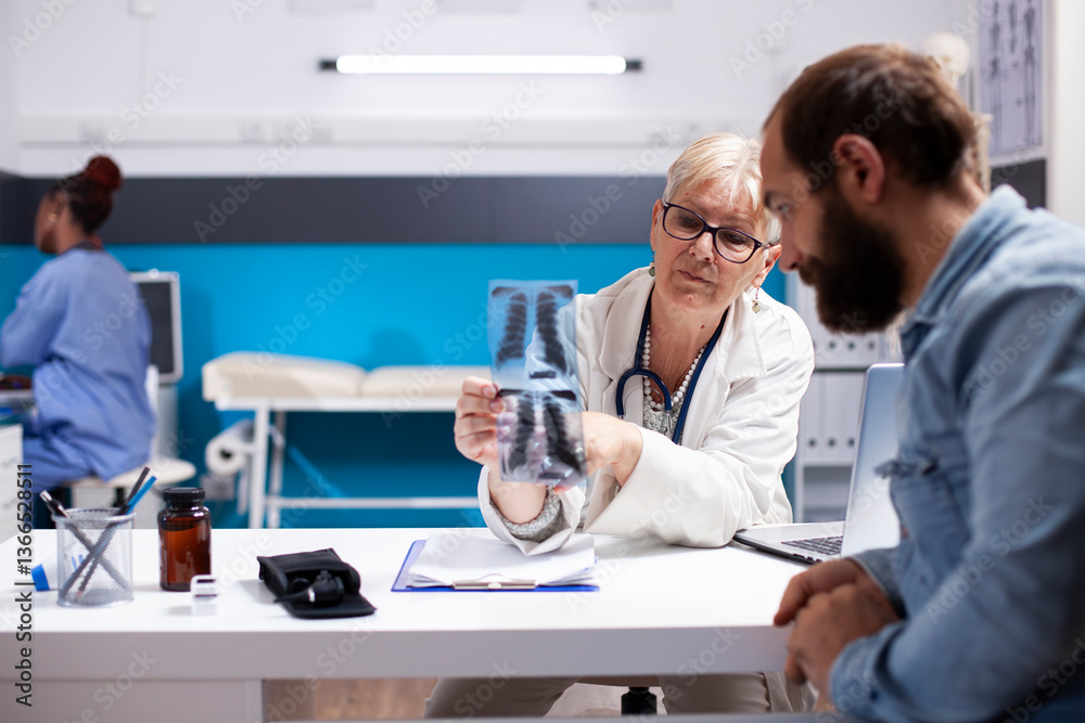 In clinic, elderly female health specialist holds xray, pointing out diagnosis details for young man with beard. Senior doctor advices male patient regarding radiology results and medical next steps.