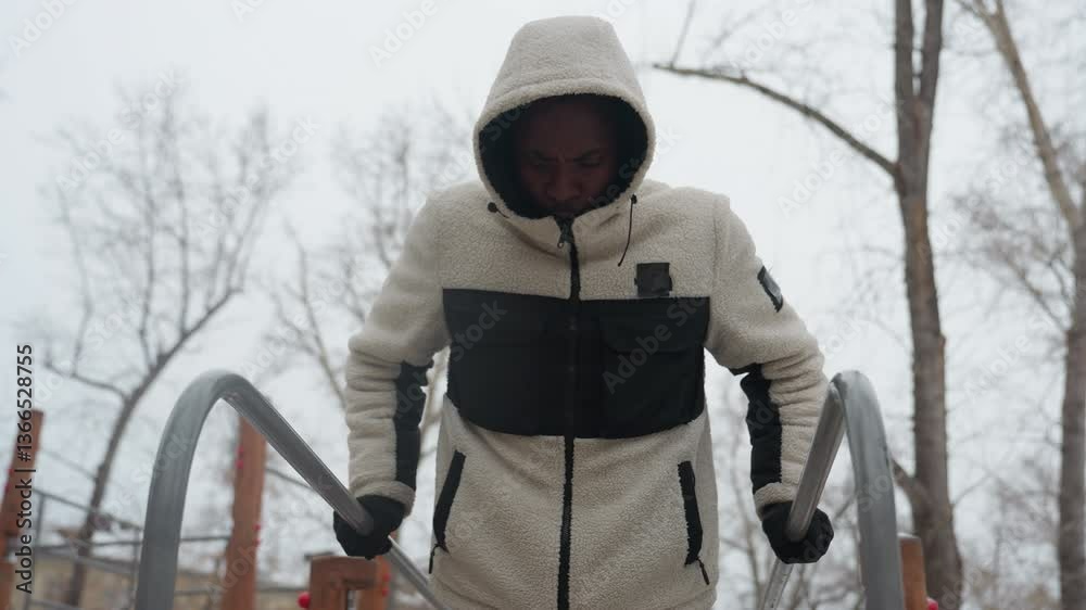 Close-up of young man performing push-up exercise for muscle buildup on outdoor parallel bars during winter, he is wearing a hooded jacket and gloves, with visible breath in the cold air