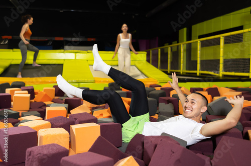 Obraz na płótnie Cheerful young man enjoying soft landing in foam pit at trampoline park, with friends jumping around w salonie