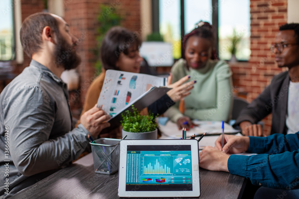 Tablet with financial charts displayed on desk as team members in the background discuss business strategies. Closeup of analytics data on device as coworkers share ideas during meeting in office.