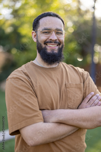 A cheerful bearded man stands confidently outdoors, smiling warmly against a vibrant backdrop. His demeanor exudes positivity and joy, inspiring those around him with a strong sense of community