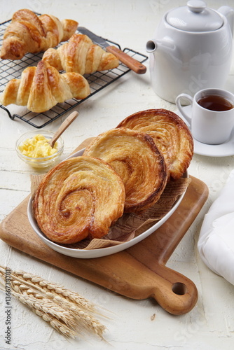 A delicious breakfast spread featuring swirl pastries and croissants, served with a cup of tea, butter, and a white teapot on a rustic wooden board, creating a warm and cozy morning vibe.