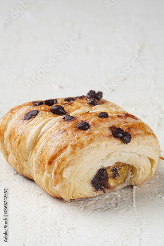 Close-up of a chocolate chip pastry with a visible banana filling, placed on a white textured surface. The pastry has a golden, flaky crust topped with chocolate chips.