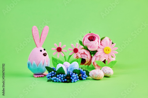 cute toy pink bunny getting ready for the easter holiday with colorful eggs and flowers on a green background