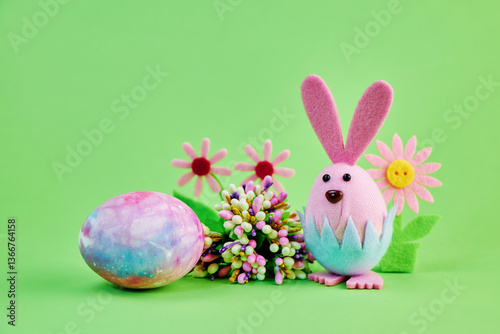 kind toy pink bunny celebrates the holiday of Easter with a large colorful egg and flowers on a green background