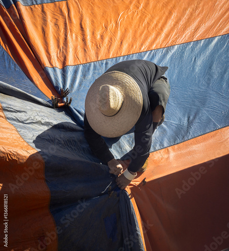Eye level with a worker wearing a straw hat installing an orange and blue  fumigation tent on a house