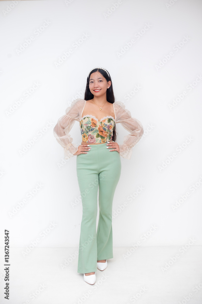 Obraz premium Young Asian woman in floral bustier and pastel green pants smiling with hands on waist against white background