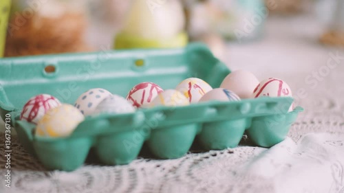 A pastel-colored carton of hand-painted Easter eggs with abstract patterns in red, blue, and yellow, placed on a lace-covered table with a softly blurred background.