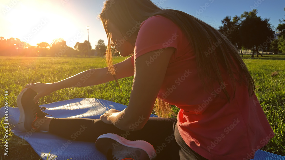 Young woman exercising on a sports yoga mat on the grass in a park during summertime sunset time.