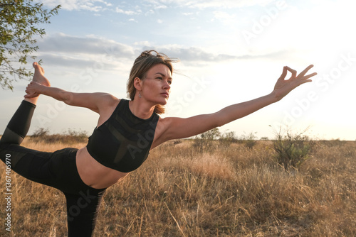 A serene young woman adopts a sunlit yoga pose against a blue sky.