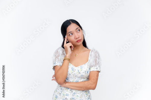 Unsure young Southeast Asian woman thinking with hand on temple feeling uncertain while in white floral dress on studio background