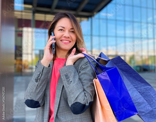 Portrait of joyful adult beautiful woman holding many paper bags with bought stuff and smiling while speaking on smartphone outdoor in city. Caucasian casual female on shopping