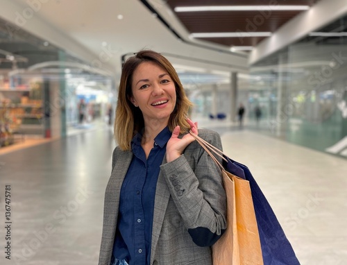 One cheerful middle-aged Caucasian woman with happy face standing in shopping mall and smiling looking at camera. Beautiful happy female with paper bags contented after shopping photo