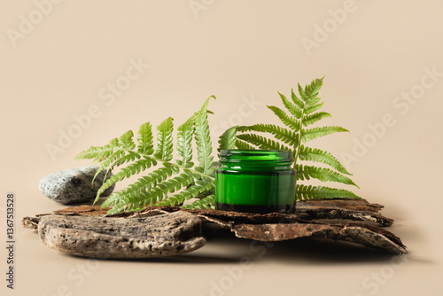 Concept of spa or moisturizer face cream in green glass jar with natural ingredients decorated fern leaves and stone on wood bark and beige background. Close up.
