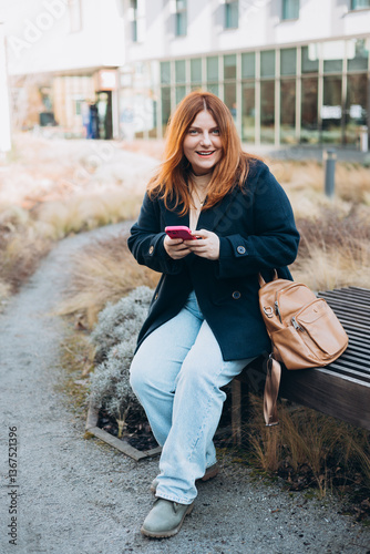 Happy woman sitting on the bench and chatting on phone outdoors. Urban, people concept. Smiling 30s redhead woman text messaging on smart phone at sunny day on the city street, spring time