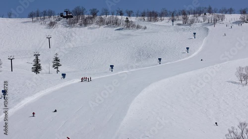 Snowboarders descending steep, wide slope on sunny day. Zoom shot
