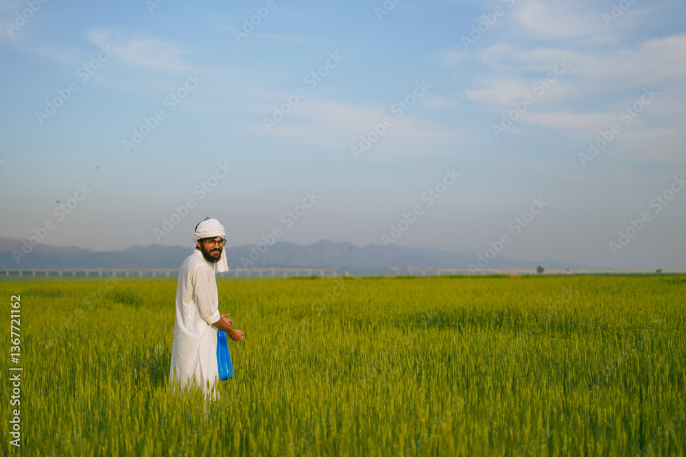 A smiling farmer wearing traditional white clothing and a headscarf stands in a lush green wheat field with mountains in the background. A smiling farmer wearing traditional white clothing and a headscarf stands in a lush green wheat field with mountains in the background.