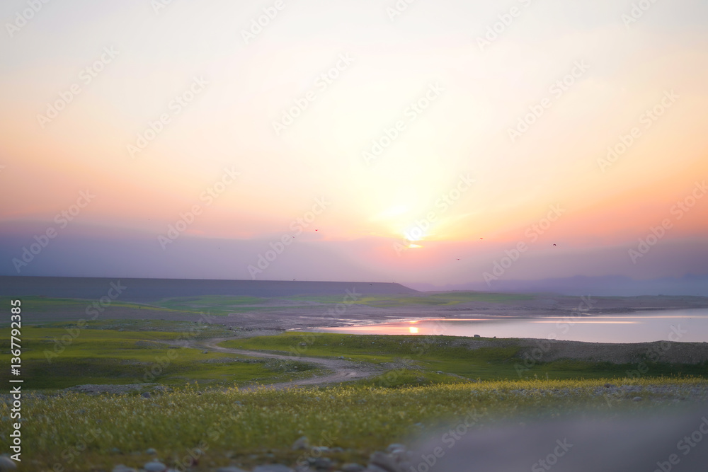 Obraz premium A breathtaking sunset over lush green fields, a winding dirt path, and a calm reflective lake, creating a peaceful and scenic view.