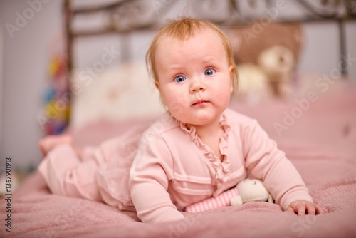 Baby girl with light hair and blue eyes lying on pink bed. Soft lighting and bedroom setting create warm and peaceful atmosphere