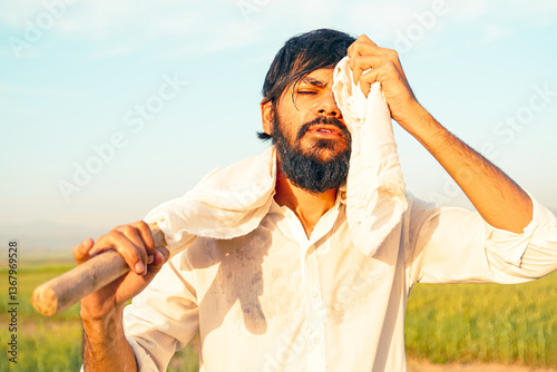 Fotografie A tired Pakistani Indian farmer wipes sweat from his face with a cloth while holding a hoe, working under the scorching sun