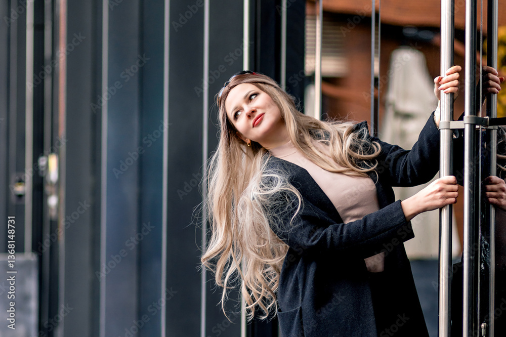 Portrait of blonde stylish woman posing near glass doors wearing black pants and coat