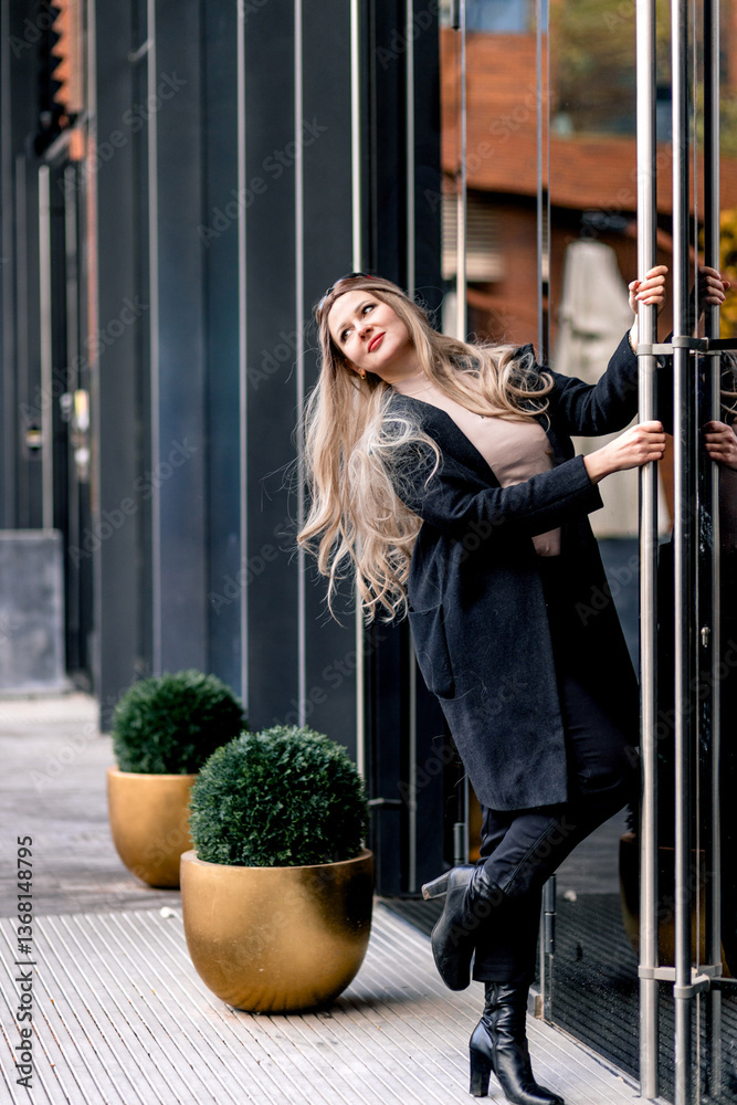 Portrait of blonde stylish woman posing near glass doors wearing black pants and coat