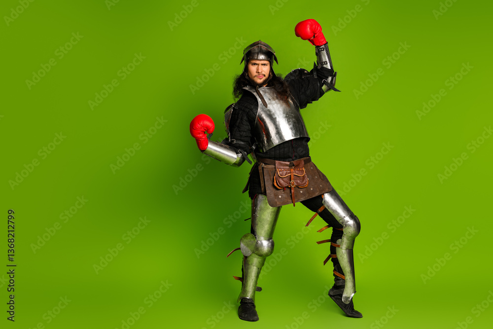 Fototapeta premium Young man dressed as medieval knight with boxing gloves in a humorous representation of past and modern era, isolated on a green background