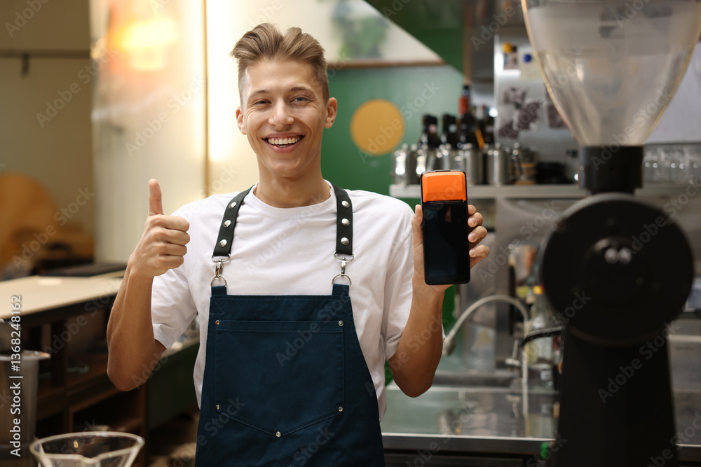 Smiling cafe worker with payment terminal showing thumbs up indoors
