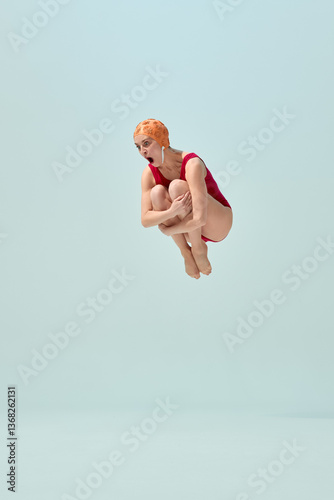 Diver in red vintage swimsuit and orange cap making cannonball pose in air with comic expression. Concept of summer fun, playful holiday, vintage swimsuit style, humor, retro sport. photo