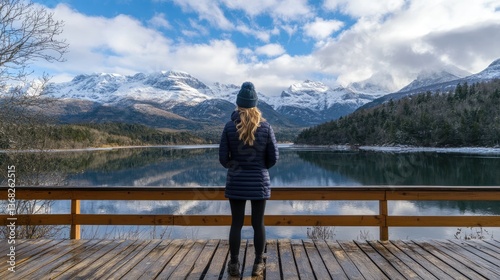 A woman stands on a wooden dock overlooking a serene lake surrounded by snow-capped mountains under a blue sky