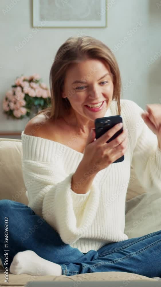 Young woman is sitting on a comfortable sofa in her living room, using her phone. She receives good news and celebrates with joy Young woman is sitting on a comfortable sofa in her living room, using her phone. She receives good news and celebrates with joy